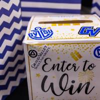 "Enter to Win" Raffle box decorated with GVSU stickers and three blue and white striped bags